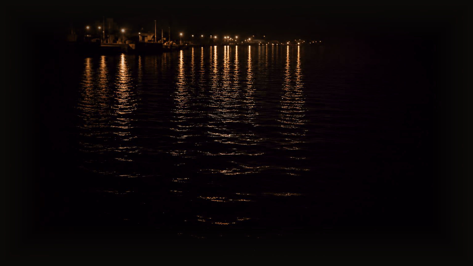 Harbor lights at dusk reflecting in long copper streaks across dark rippling water, dissolving into darkness